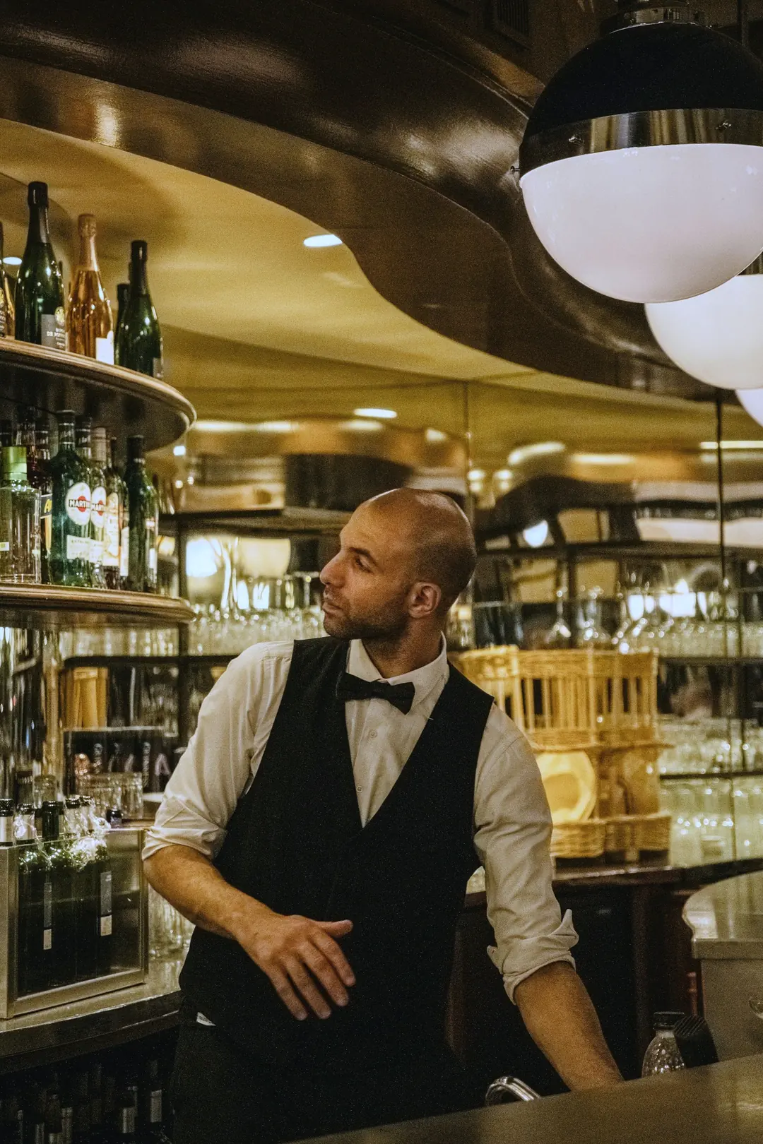 Bartender in a black waistcoat and bow tie behind a brass and glass bar