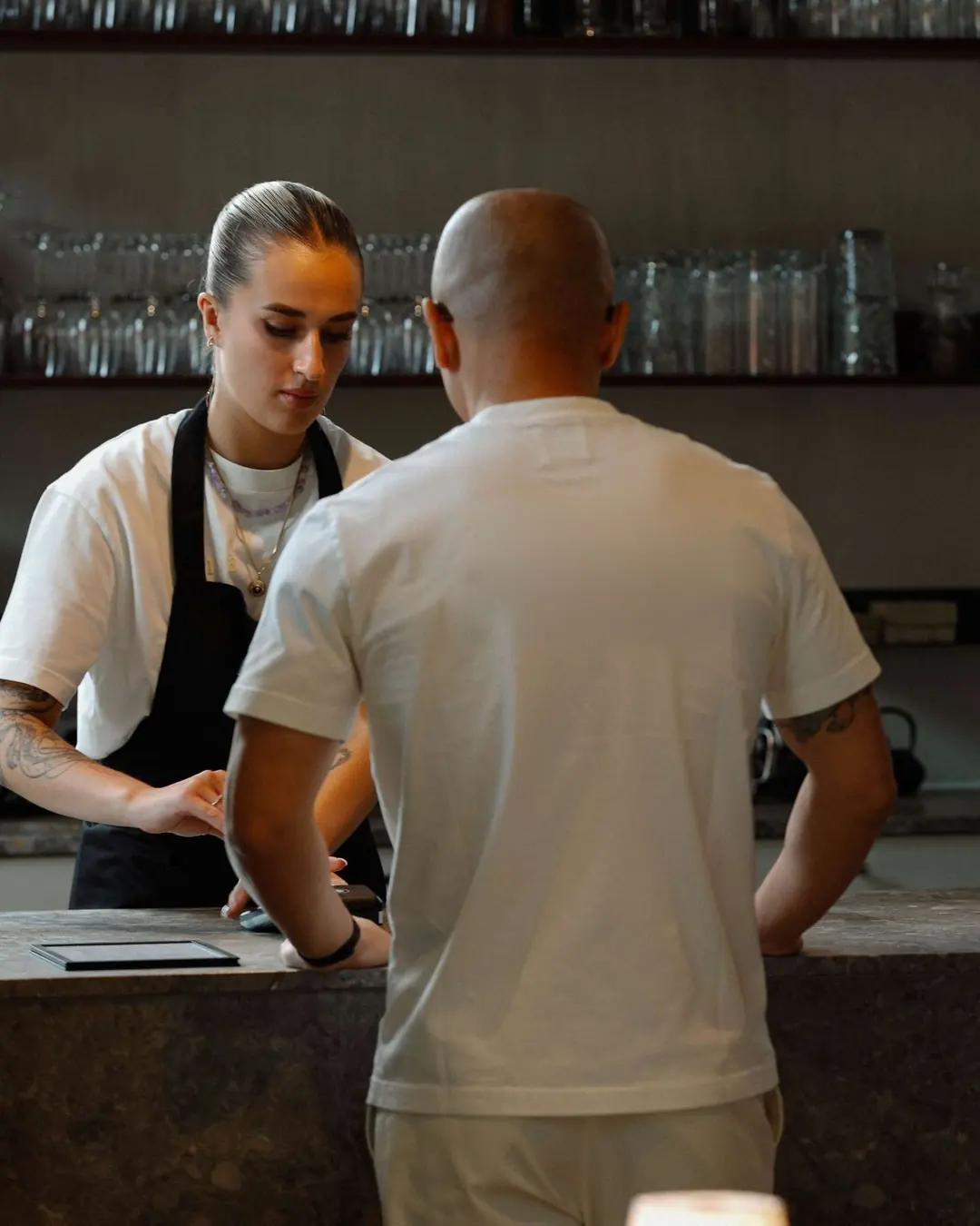 Two hotel team members at the bar in cream T-shirts, one with a black apron