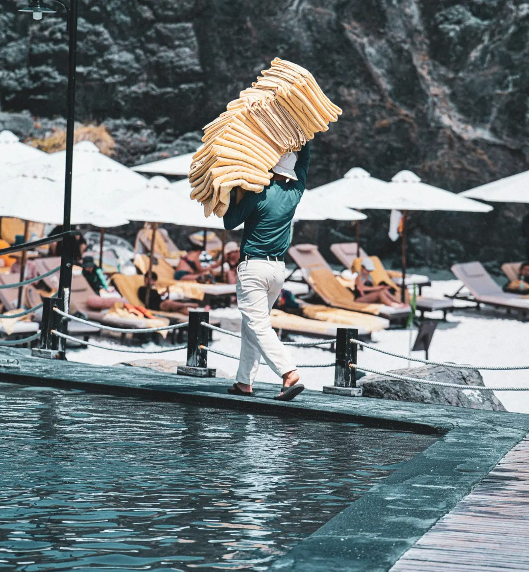 Pool attendant carrying a tall stack of folded towels along a resort pool deck