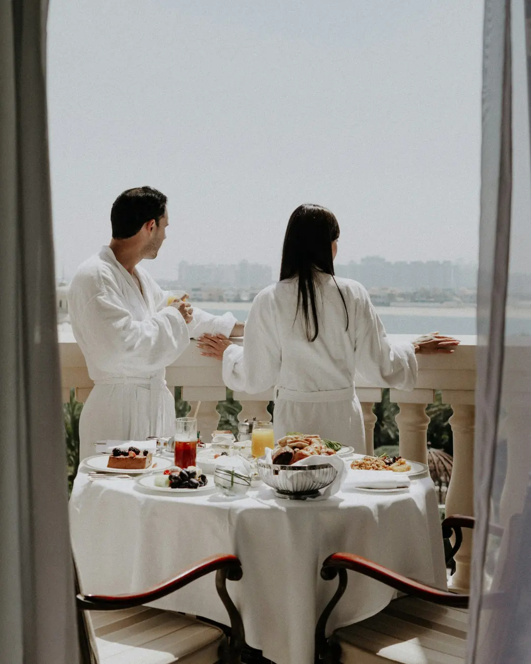 Two guests in white bathrobes at a laid breakfast table on a hotel balcony above the water