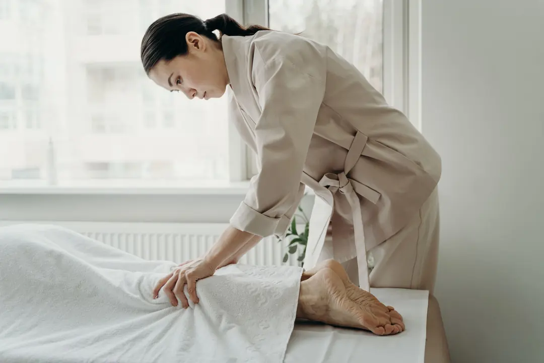 Spa therapist in a belted beige wrap tunic smoothing a white sheet on a treatment bed by a window
