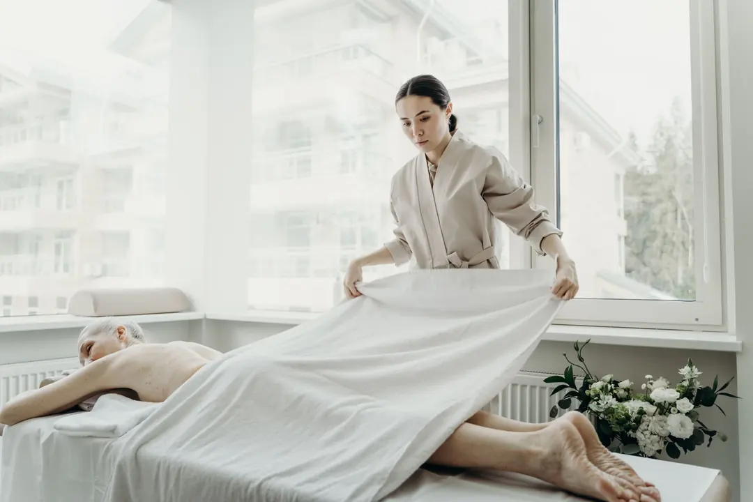 Therapist in a beige wrap tunic laying a white sheet over a guest on a treatment bed in a bright room