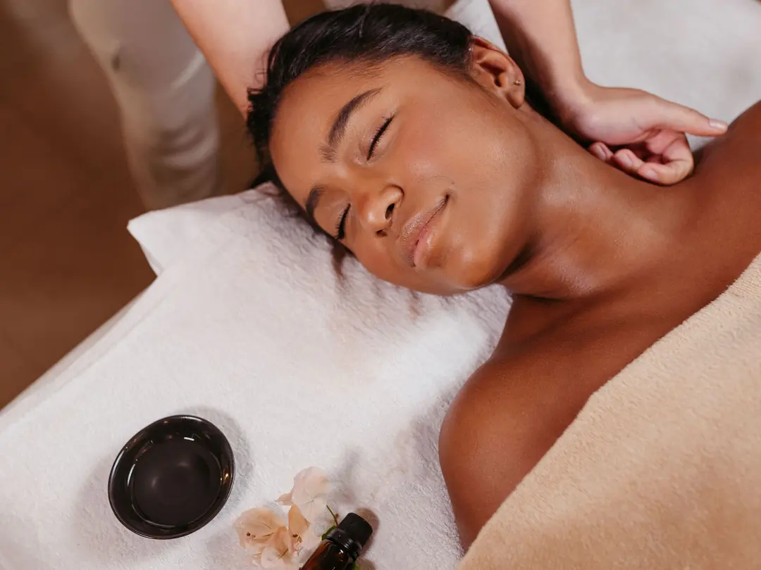 Guest resting on white towelling under a sand-coloured terry towel during a neck massage, a bowl and oil beside her