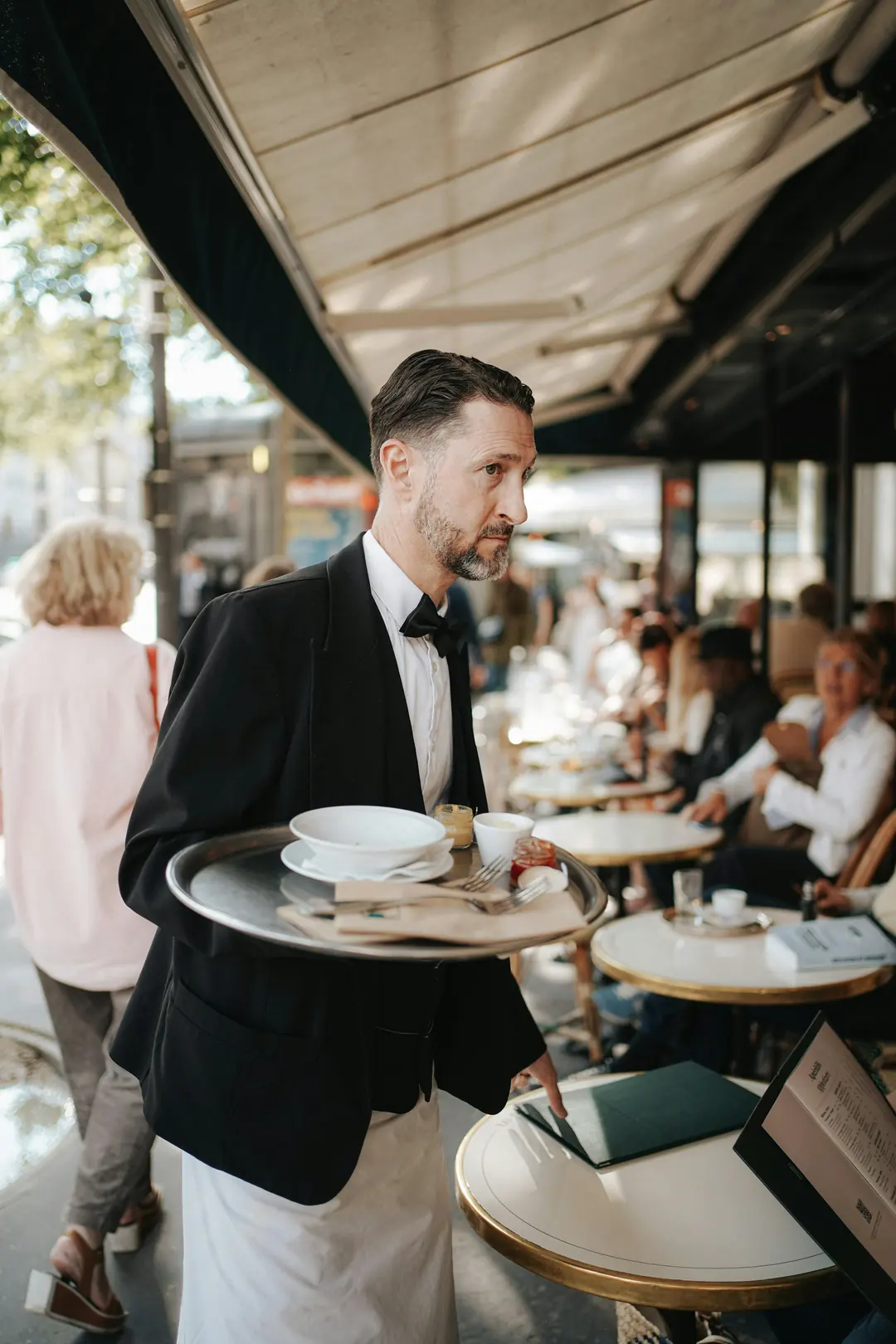 Waiter in a black jacket and long white apron carrying a tray between café tables